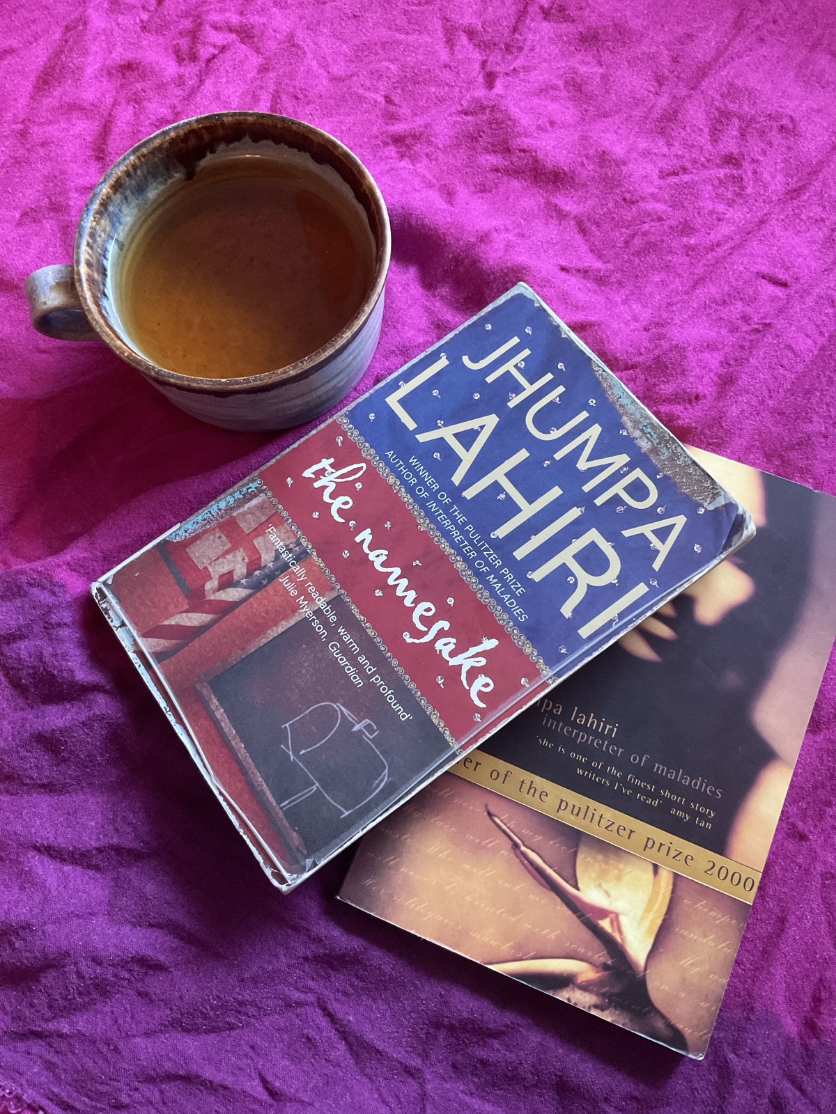 The Namesake and Interpreter of Maladies by Jhumpa Lahiri are lying on a pink fabric that fades into purple towards the lower end of the picture. The books overlap. The Namesake is on top of Interpreter of Maladies covering the latter's left top. To the left of the books is a blue-brown cup filled with a brown-coloured tea.